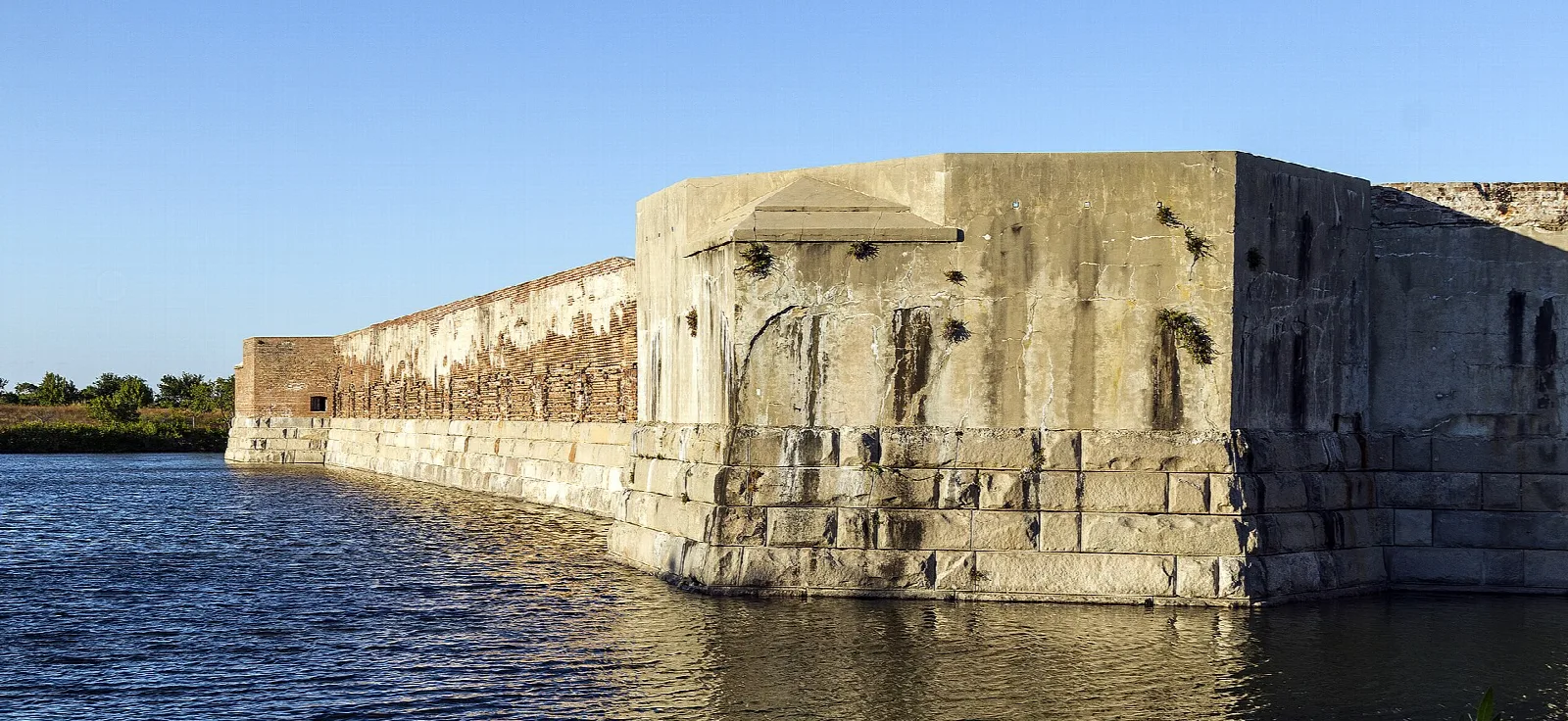 Weathered fort wall rising from water with a long wall beyond