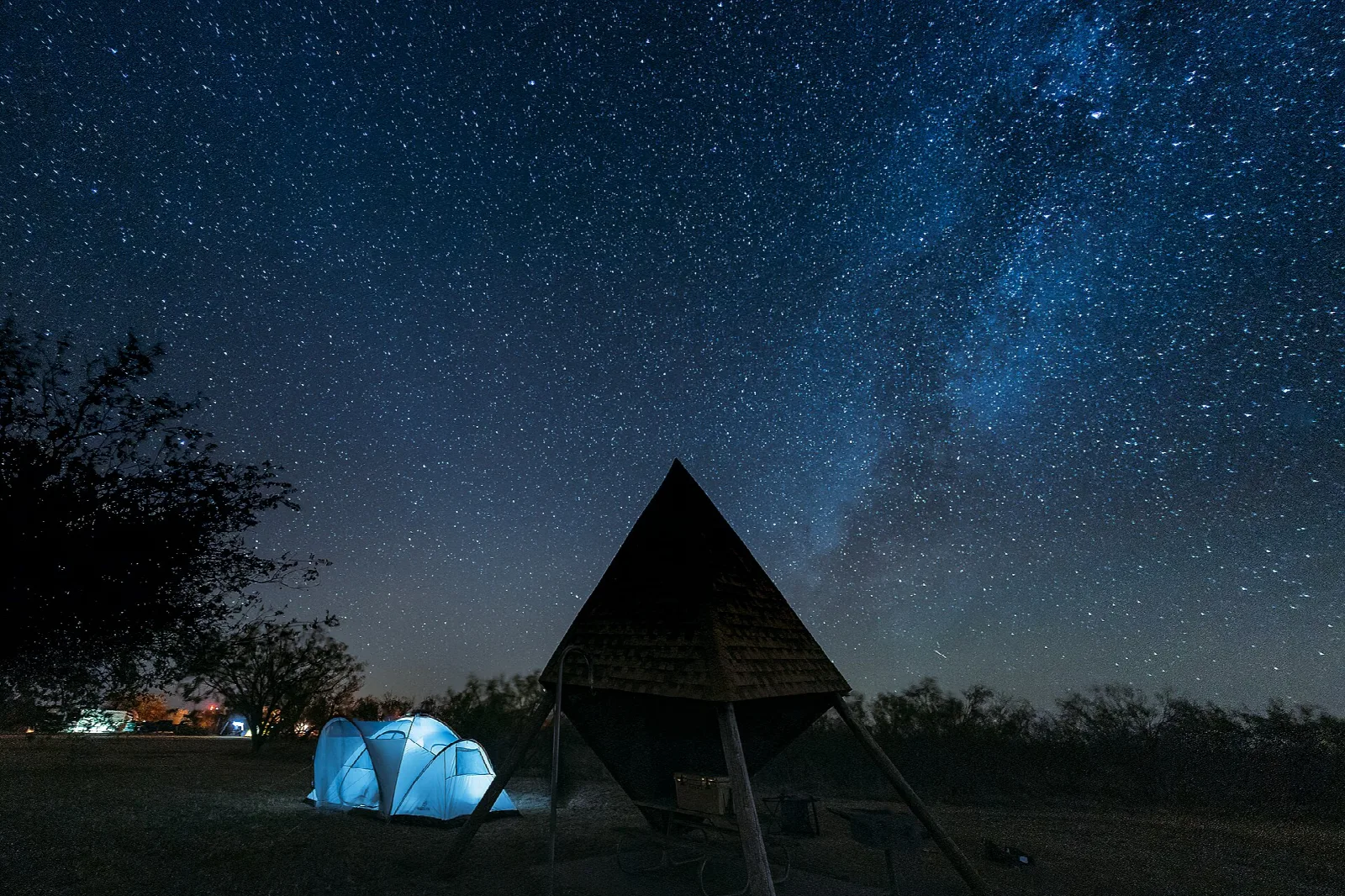 Milky Way over a pyramid-roofed shelter and a lit tent at night