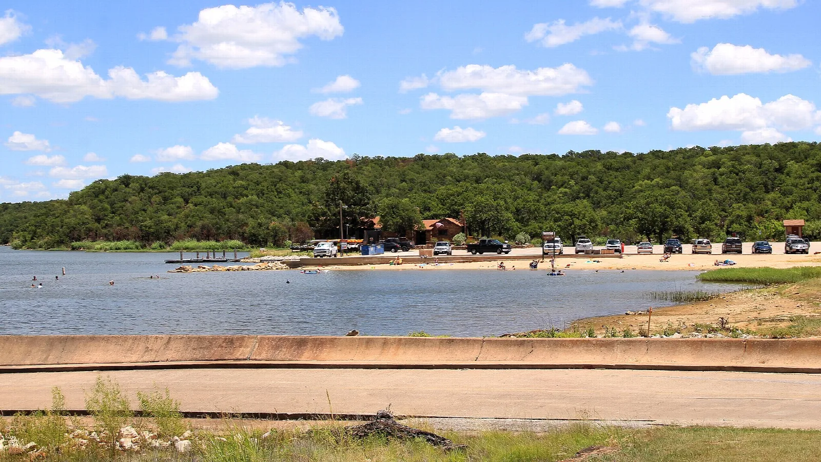 Sandy swimming area on the lake with cars and trees behind