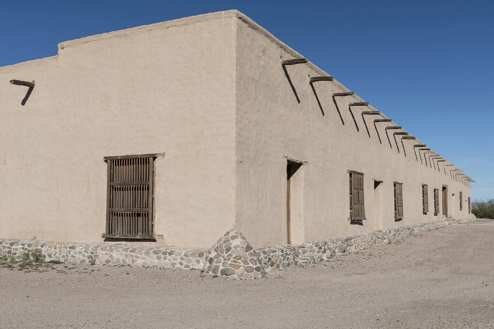Long tan adobe building with wooden beams and a shuttered window under a blue sky