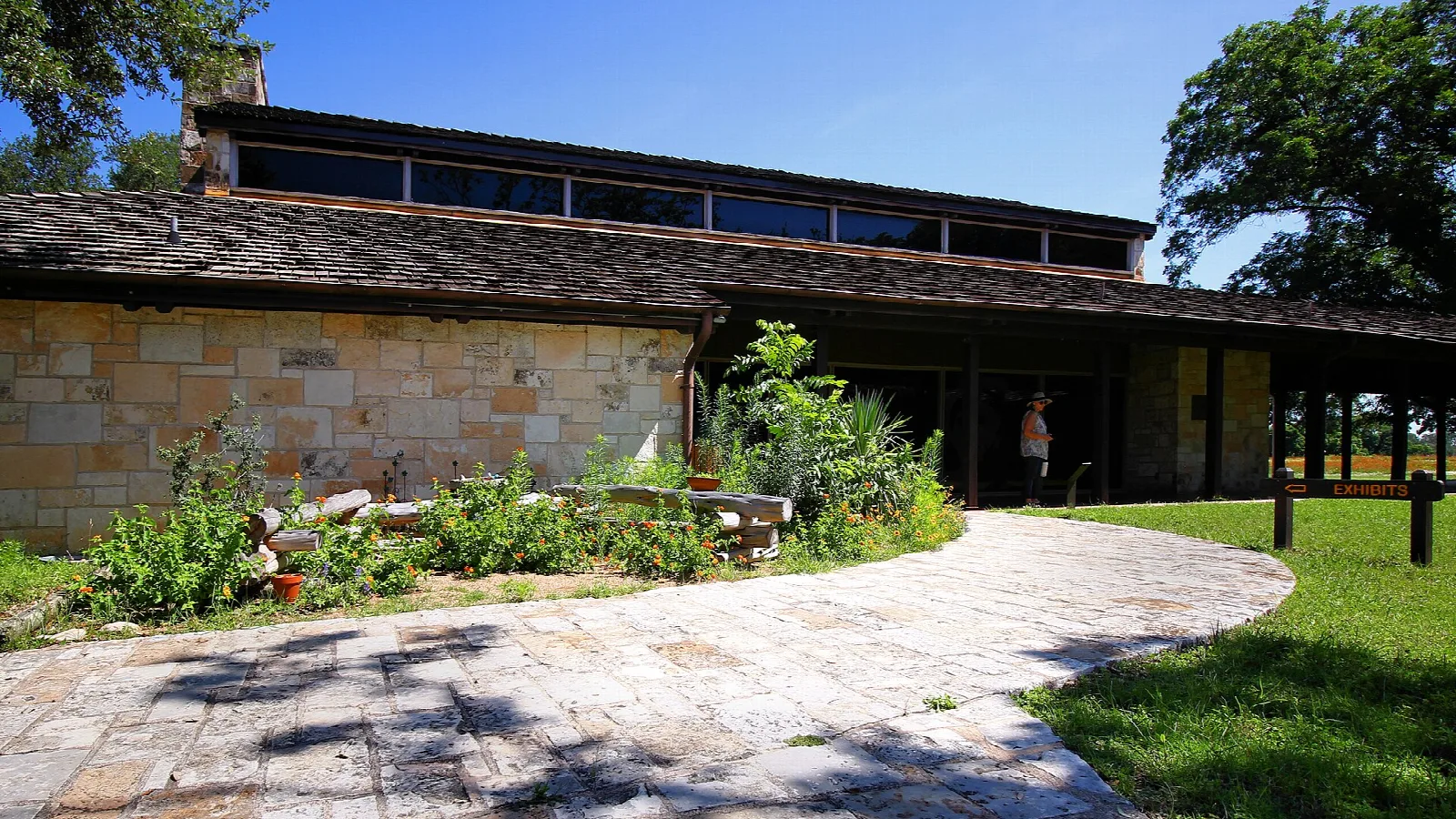 Low stone and wood exhibits hall with a paved walkway and plants