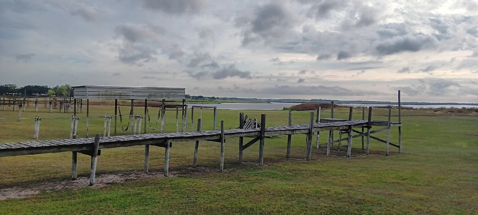 Wooden walkway over grass beside a wide lake under cloudy sky