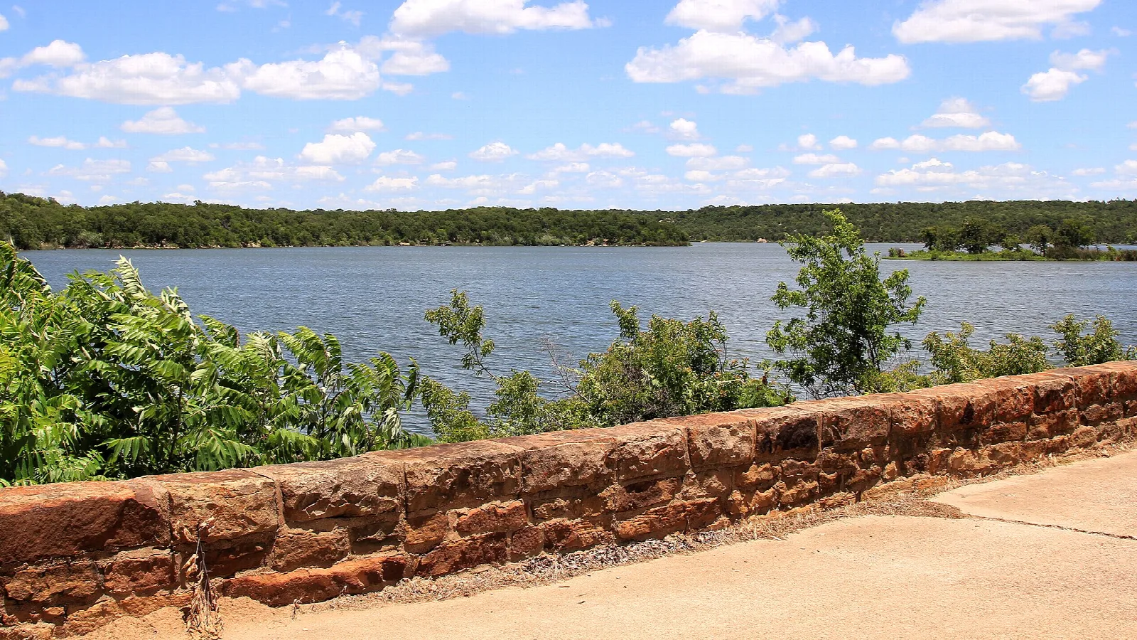 Red stone wall beside a path with a lake and trees behind