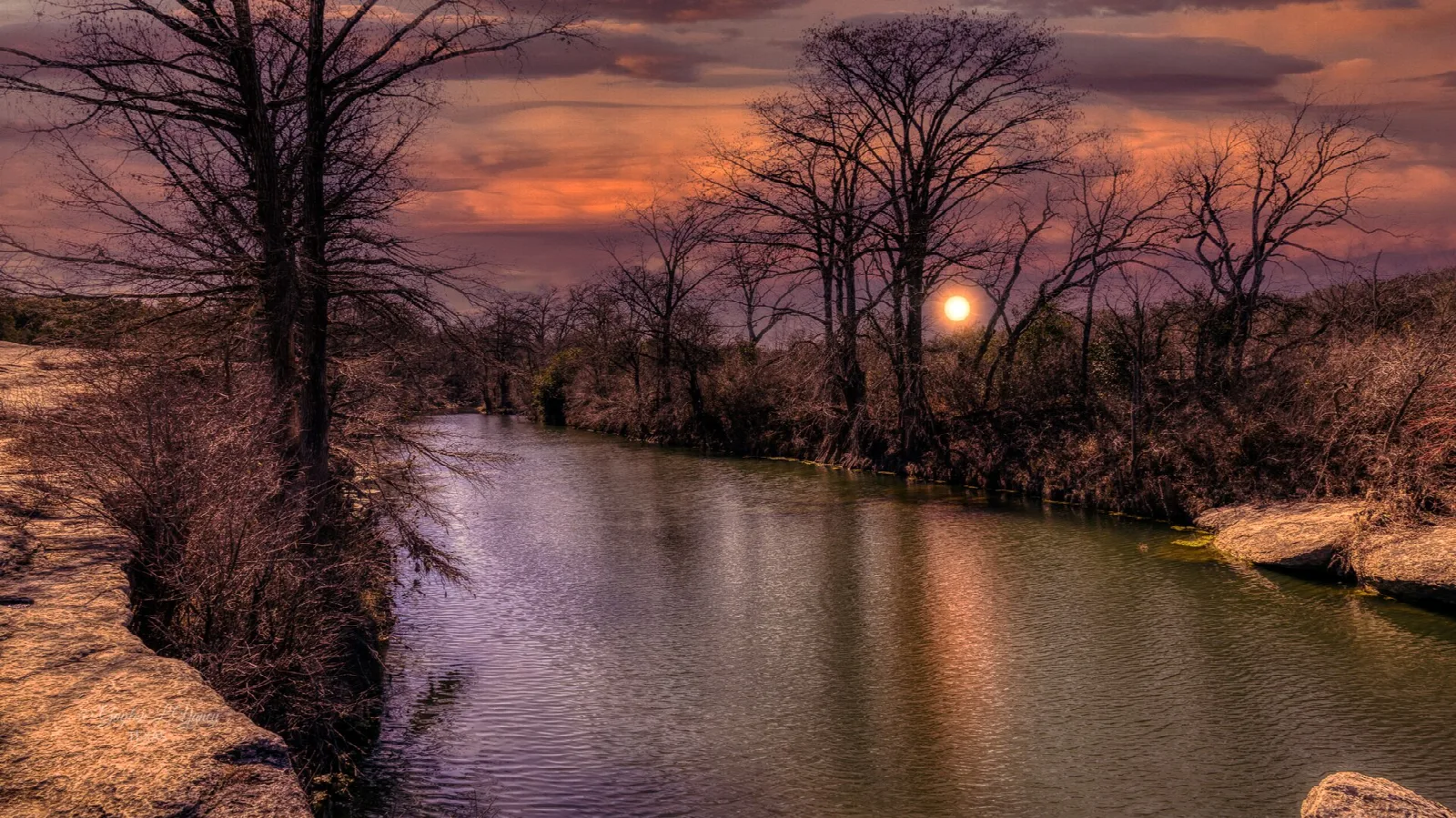 Calm creek at sunset between rocky banks and bare trees
