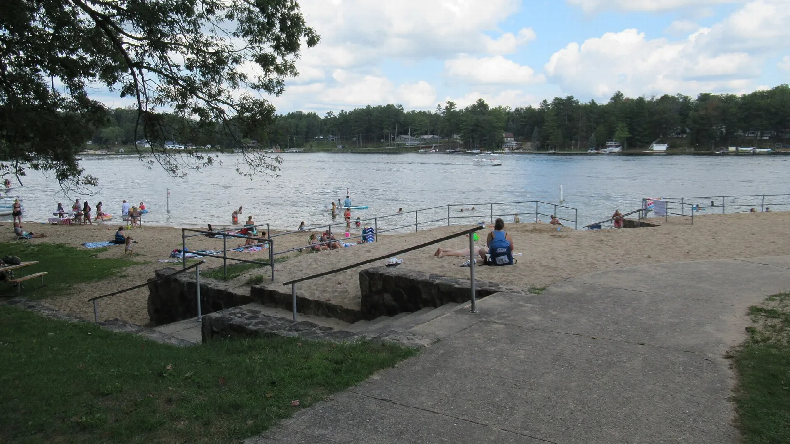 Sandy swim beach with people at the water's edge on a lake lined with trees