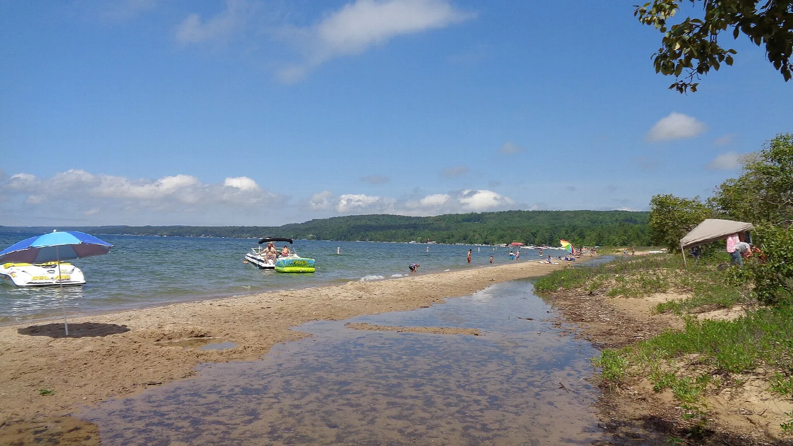 Sandy shore with shallow water, boats offshore and wooded hills across the lake