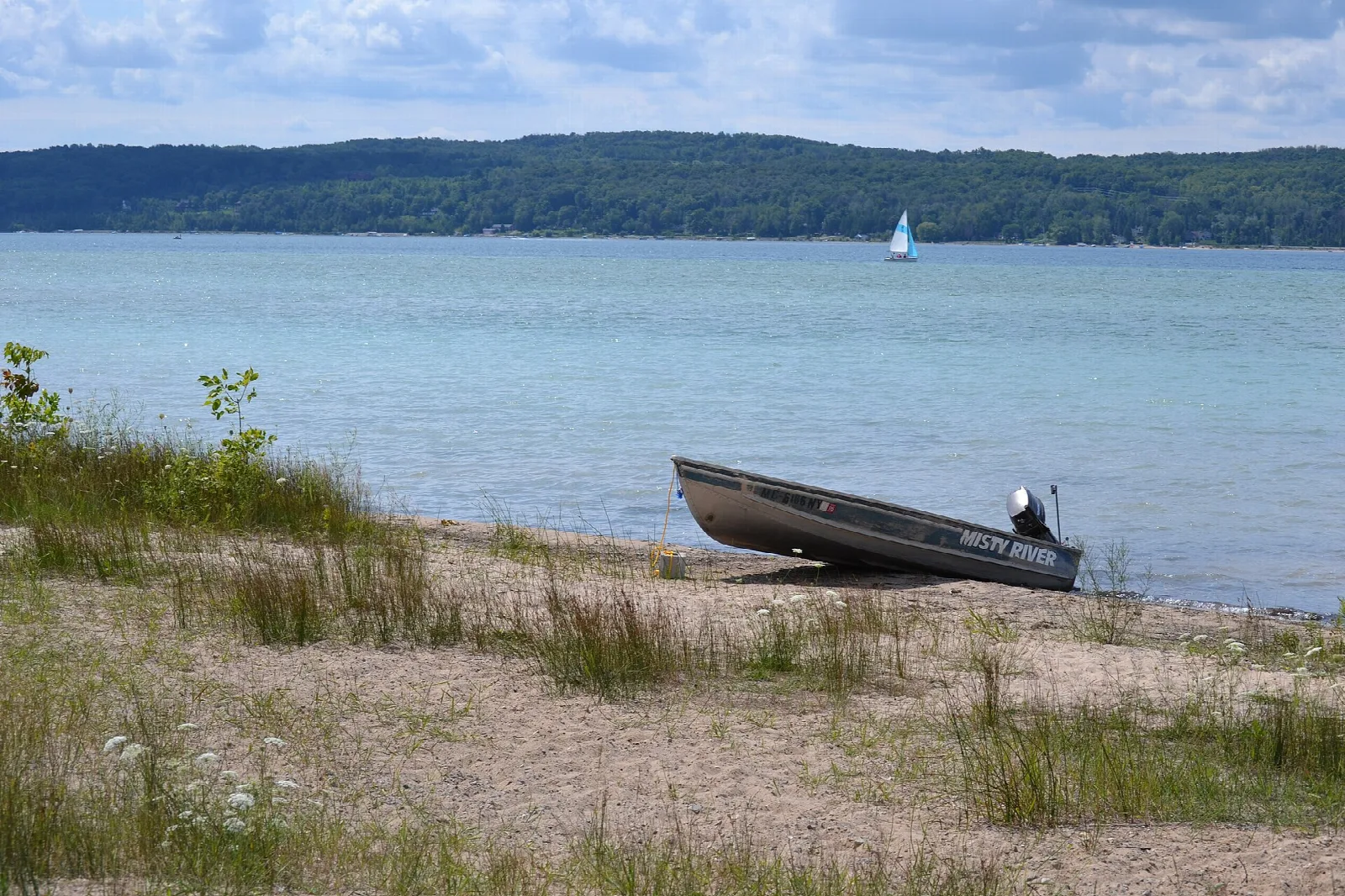 Small boat pulled up on a grassy sand shore with a sailboat on the lake beyond