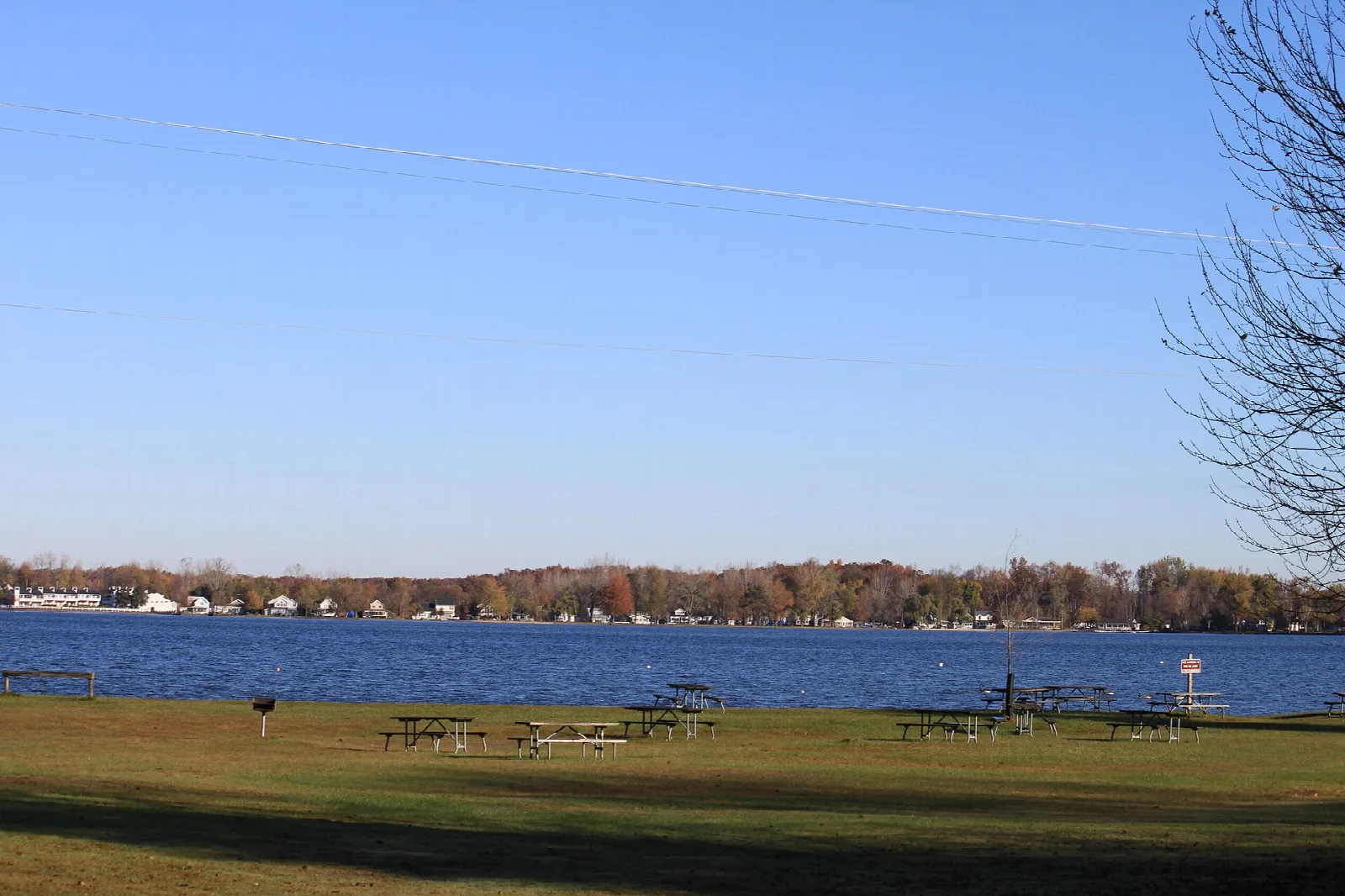 Grassy picnic area with tables beside a blue lake and houses on the far shore