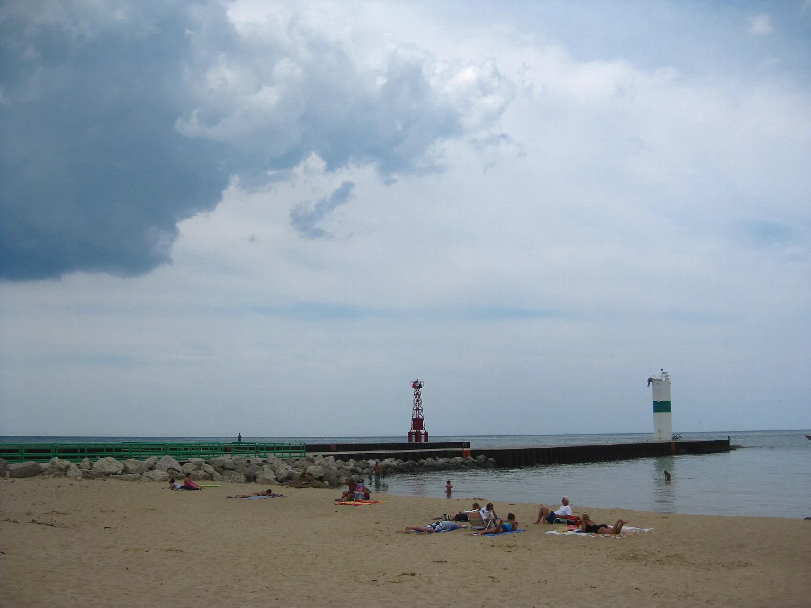 Sandy beach with people near a long pier and two harbor lights under cloud