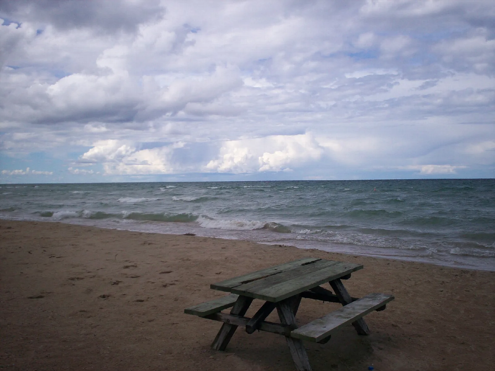 Picnic table on a sandy beach beside wind-rippled lake water under cloudy sky
