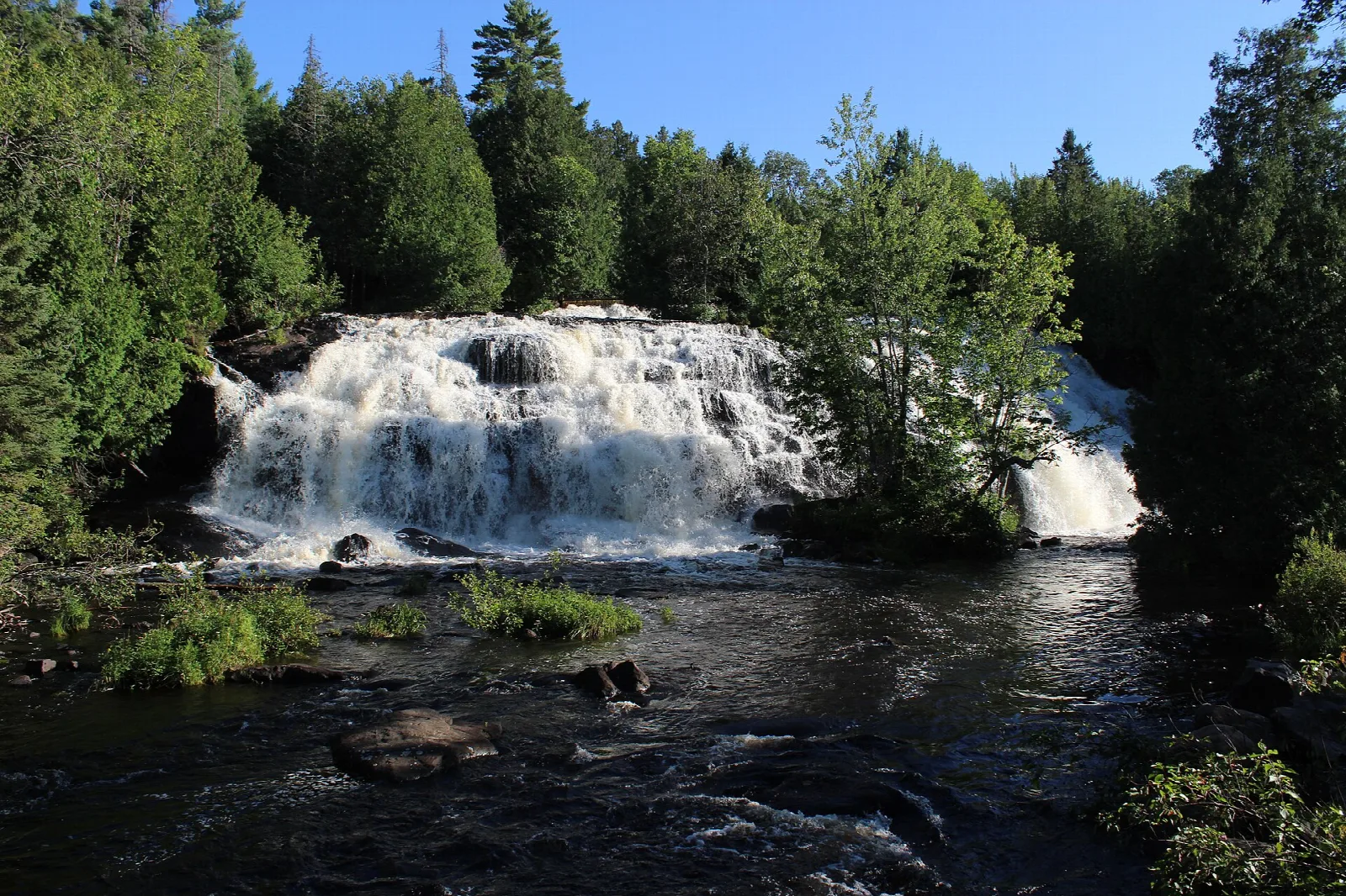 Waterfall spreading across rocks above a rushing river with evergreens behind