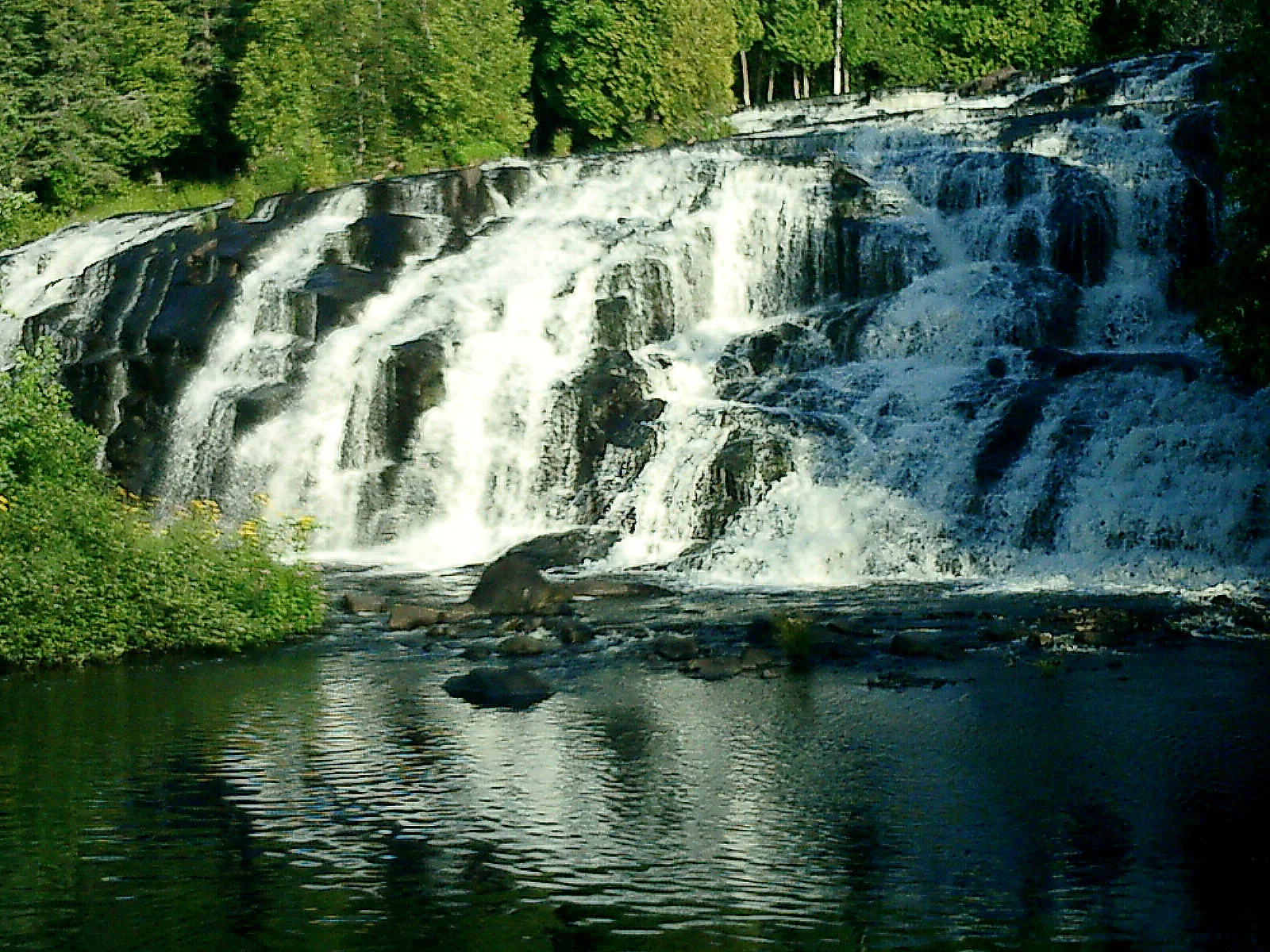 Wide tiered waterfall dropping into a dark pool with forest above