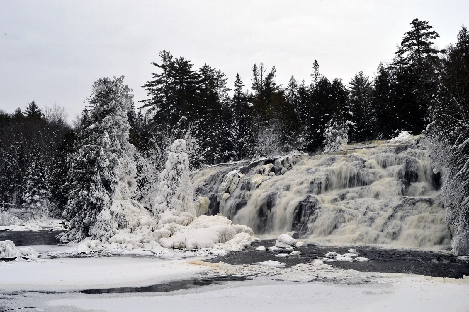 Snow-covered evergreens beside a frozen waterfall with ice sculptures