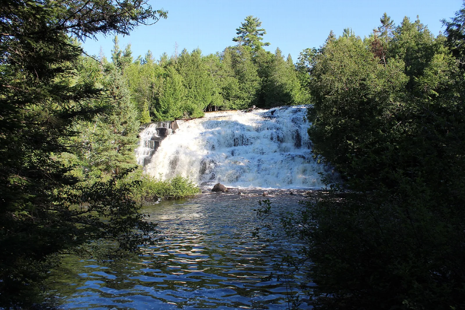 Broad waterfall seen across the river through evergreen trees under blue sky