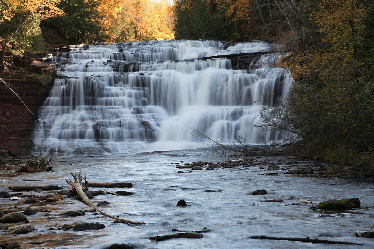 Wide, many-stepped waterfall over rock ledges with autumn trees at left