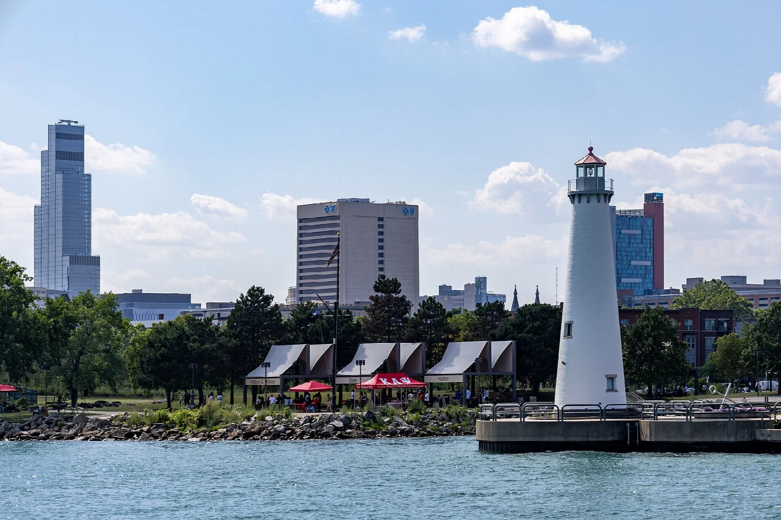 White lighthouse beside a row of covered shelters with downtown towers behind