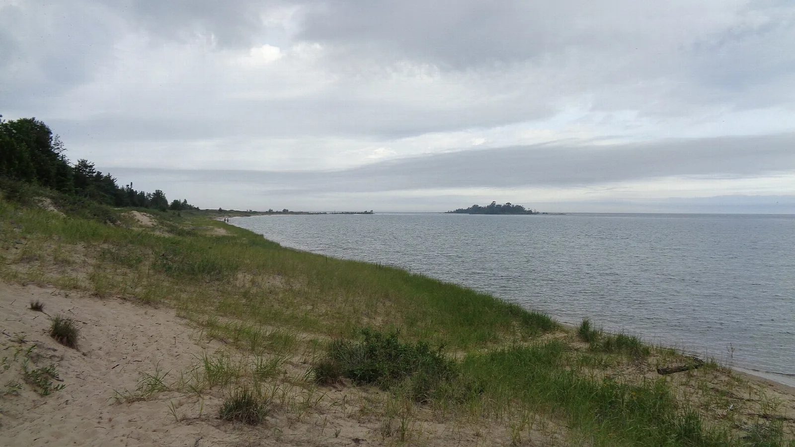 Grassy dune slope above a calm gray lake with a small island in the distance