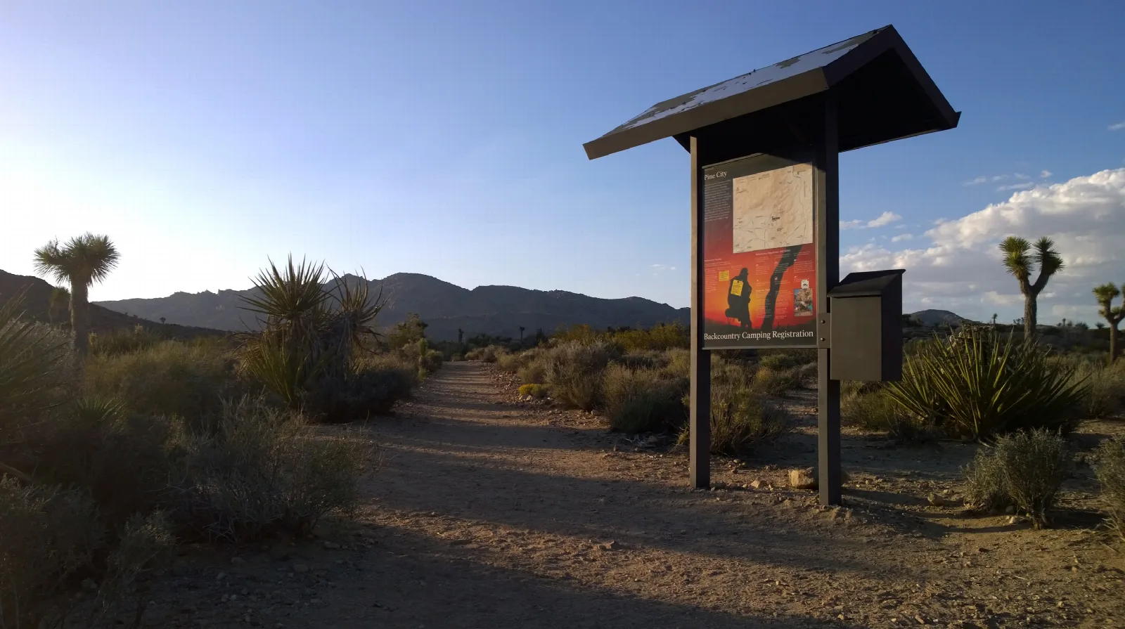 A backcountry board with hiking and backpacking info next to a dirt trail heading towards mountains.