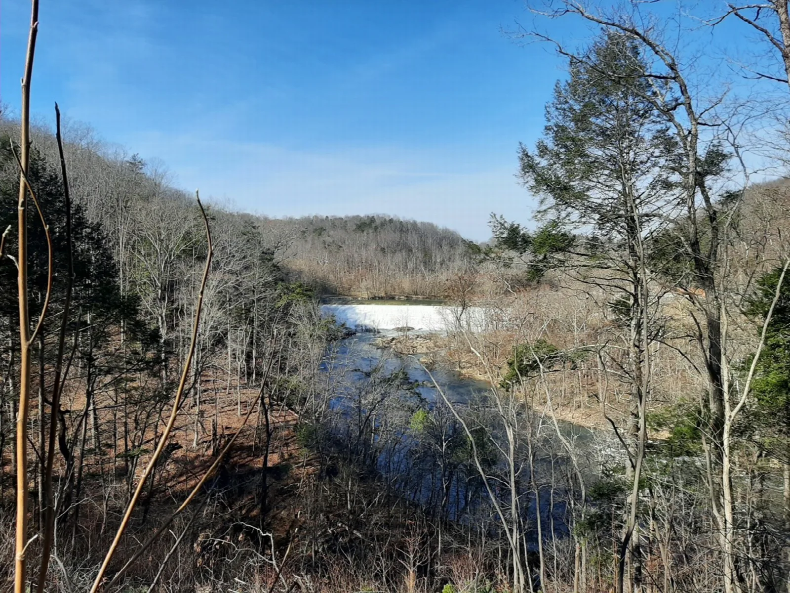 A low, white dam on a river surrounded by winter-bare forested hills