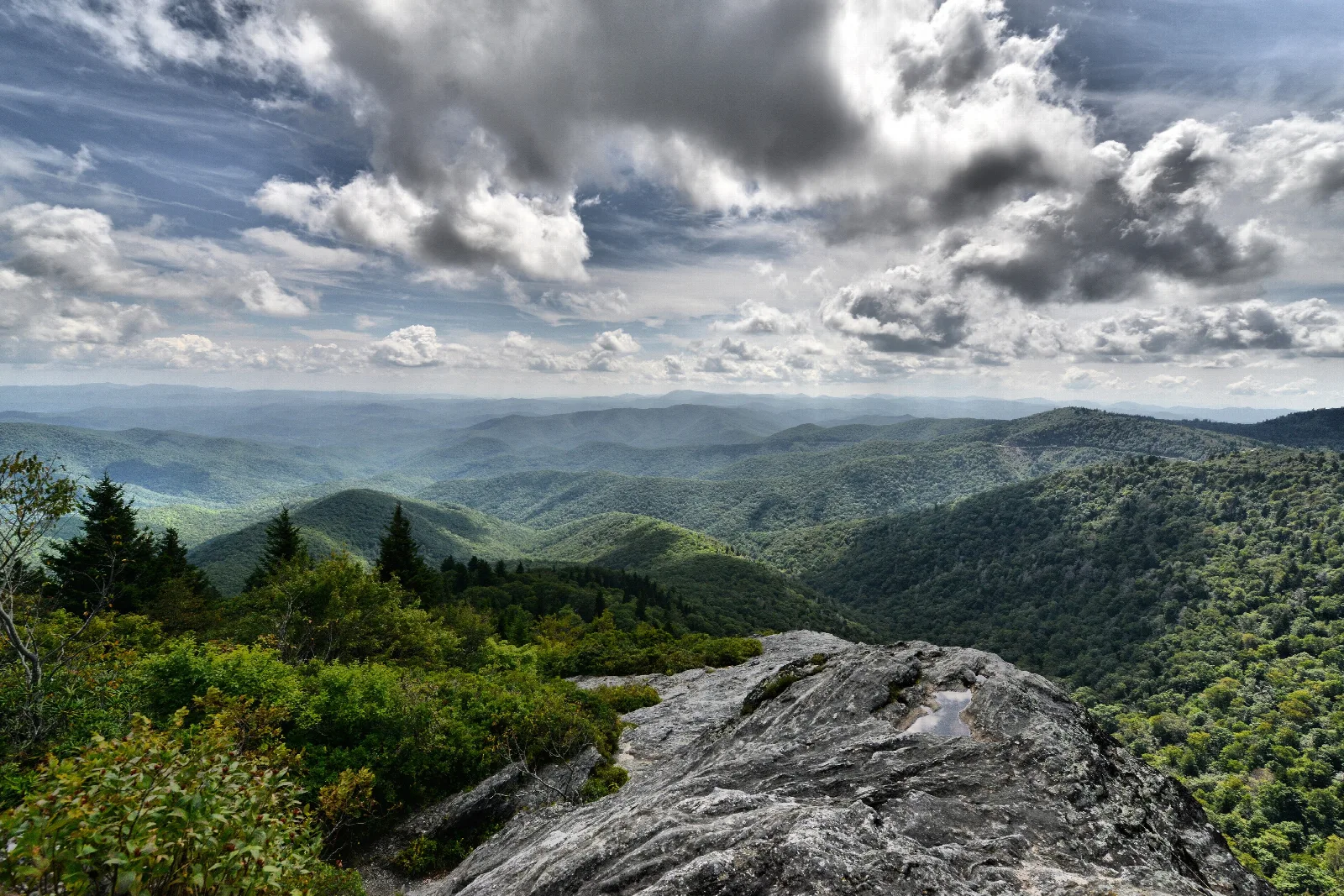 A rocky outcrop looks out over green layered mountains and white clouds