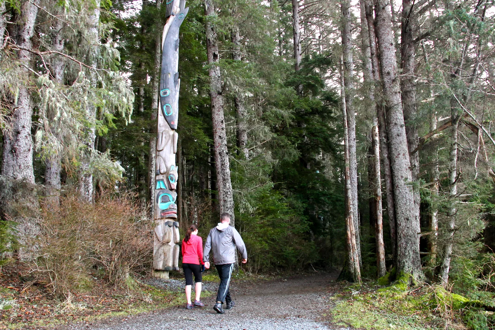 A man and woman walk by trees and a totem pole on a gravel path